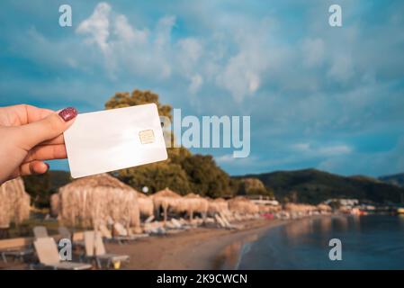 Weiße Bankkarte in Frauenhand auf Hintergrund des Strandes mit Sonnenliegen und Sonnenschirmen in Moraitika, Korfu, Griechenland. Das Konzept Der Bezahlung Für Entspannung Stockfoto