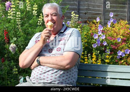 Älterer Mann, der auf einer Bank sitzt und ein Eis isst. Lächelnd und in Richtung der Kamera schauend. Glücklich. Stockfoto