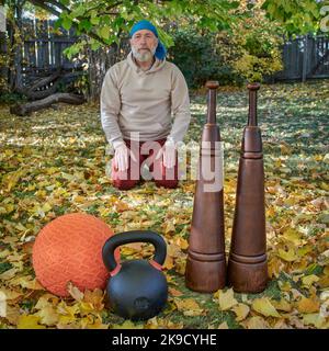 Schwere eiserne Kugelhantel, Slam Ball und hölzerne persische Keulen in einem Hinterhof mit einem älteren männlichen Seiza, der in einer Hintergrund-Herbstlandschaft sitzt Stockfoto