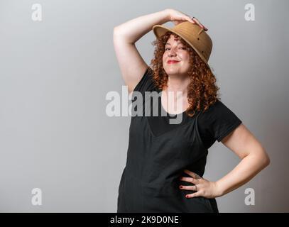 Glückliche Frau mit roten lockigen Haaren, die auf grauem Hintergrund posiert, mit Kopierraum, der einen Safarihut trägt Stockfoto