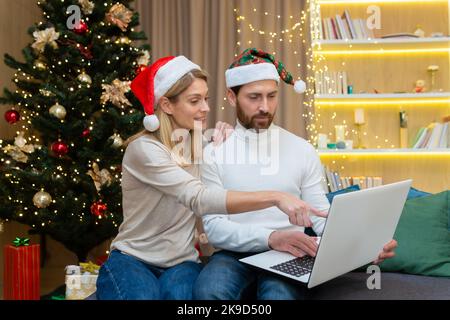 Das junge Paar, Ehemann und Ehefrau, arbeiten online zu Hause zusammen, mit einem Laptop in der Küche, glücklich zusammen, während des neuen Jahres und der Weihnachtsferien. Stockfoto