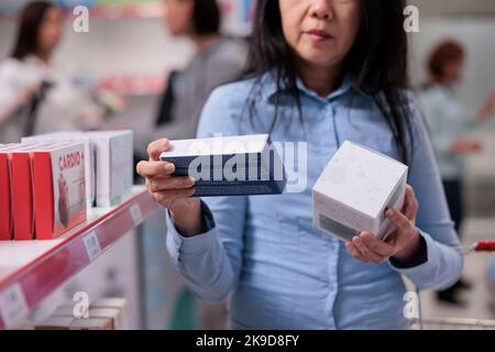 Weibliche Klientin, die sich Supplements im Apothekengeschäft ansieht, Boxen mit Vitaminen und Medikamentenflaschen in den Regalen untersucht. Drogeriekunden kaufen medizinische Produkte und Verbrauchsmaterialien. Stockfoto