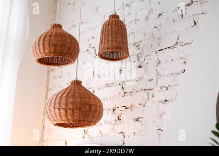 Drei Rattan-Kronleuchter in verschiedenen Formen auf weißem Backstein Wand Hintergrund. Strohlampenschirm im gemütlichen Wohnzimmer. Umweltfreundliche Innenarchitektur mit Natur Stockfoto