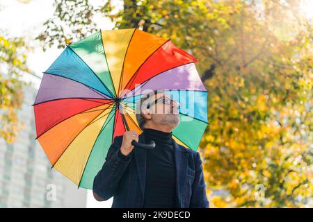 Grauhaariger Mann mittleren Alters in formellen Kleidern, der im Stadtpark herumläuft und Regenbogenschirm dabei hält, der nach oben schaut. Horizontale Außenaufnahme. Hochwertige Fotos Stockfoto