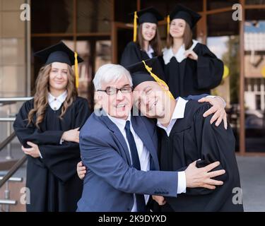 Vater und Sohn umarmen sich bei der Abschlussfeier. Der Elternteil gratuliert dem Hochschulabsolventen. Stockfoto