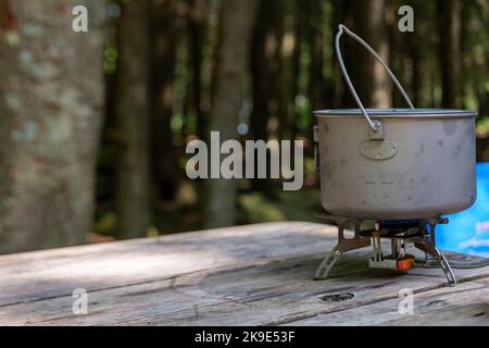 Ein Titantopf zur Herstellung von Lebensmitteln auf einem Gasherd auf einem Campingtisch aus Holz im Wald. Stockfoto