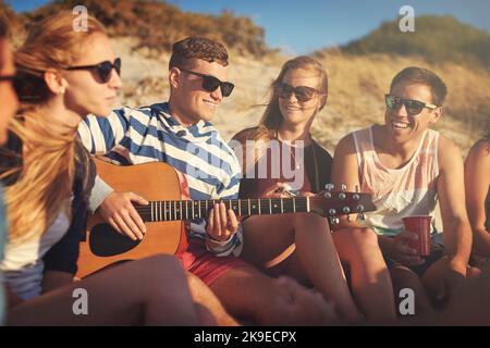 Deine Freunde sind deine größten Fans. Ein hübscher junger Mann, der an einem Sommertag am Strand für seine Freunde Gitarre spielt. Stockfoto