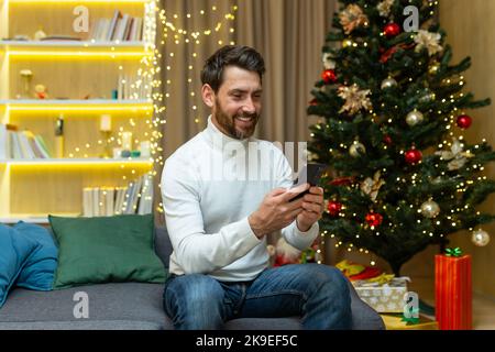 Ein junger Mann hält ein Telefon und sendet, schreibt, gibt Grußbotschaften mit Weihnachts- und Neujahrsferien an Freunde und Verwandte ein. Sitzen zu Hause mit Feiertagsdekor und Weihnachtsbaum, lächelnd. Stockfoto