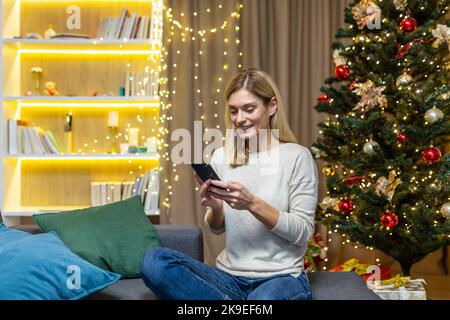 Eine glückliche junge Frau hält ein Telefon und bestellt für ein festliches Abendessen eine Lieferung. Sitzen auf dem Sofa zu Hause mit Neujahr und Weihnachtsdekorationen, Lichter und Weihnachtsbaum. Stockfoto
