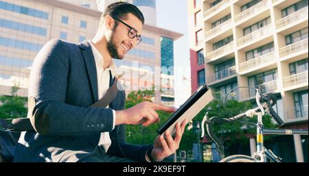 Ein hübscher junger Geschäftsmann, der sein digitales Tablet benutzt, während er sich neben sein Fahrrad in der Stadt setzte. Stockfoto