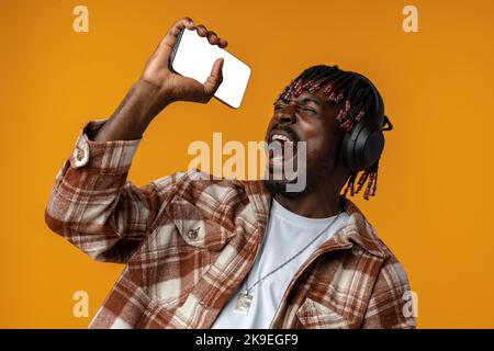 Afrikanischer Mann in Kopfhörern singt Lied, während er Musik auf dem Smartphone auf gelbem Hintergrund hört Stockfoto
