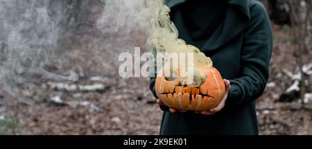 Eine gruselige Hexe hält einen dampfenden Kürbis in einen tiefen Wald. Jack o Laterne strahlt für halloween gelben Rauch aus Stockfoto