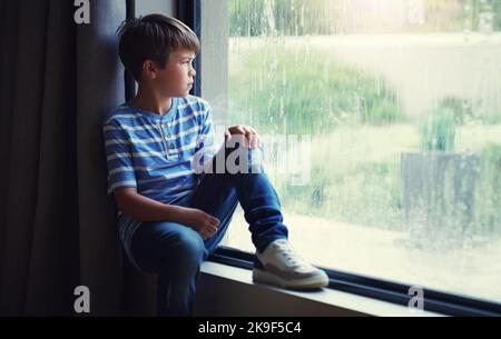 Der Regen spülte seinen Spaß für den Tag weg. Ein trauriger kleiner Junge beobachtete den Regen durch ein Fenster zu Hause. Stockfoto