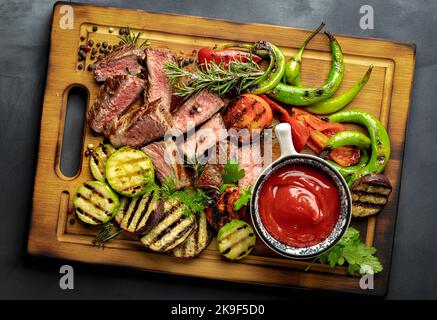 Gebratenes mittelschweres Steak mit gegrilltem Gemüse und Ketchup-Sauce auf Holzschneidebrett auf dunklem Hintergrund Stockfoto