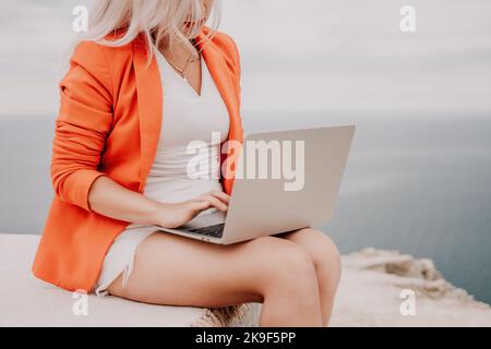 Der digitale Nomade, eine Geschäftsfrau in rotem Blazer mit einem Laptop, sitzt während des Sonnenuntergangs auf den Felsen am Meer und führt eine Geschäftstransaktion online von einem aus Stockfoto