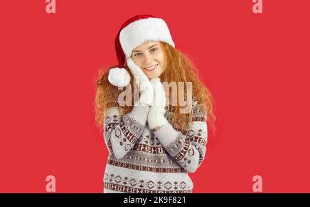 Bescheidenes rothaariges lächelndes Mädchen mit Weihnachtsmann-Hut, weißen Fäustlingen und Pullover auf rotem Hintergrund. Stockfoto