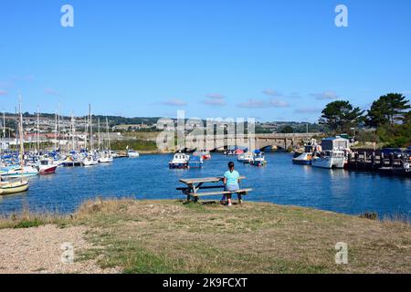 Brücke über die Axe im Hafen und eine Frau, die auf einer Picknickbank sitzt, Axmouth, Devon, Großbritannien. Stockfoto