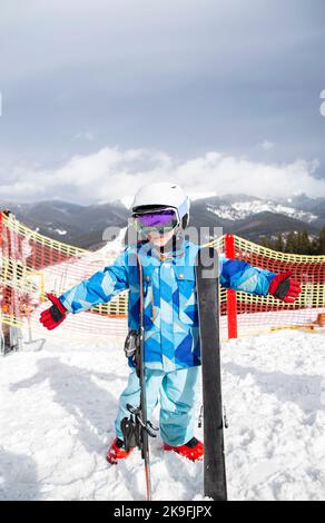 Vorschuljunge im Helm, blaue Overalls, Skischuhe steht im Schnee auf den Bergen, hält Skier. Winter aktive Unterhaltung für Kinder, Sport ed Stockfoto