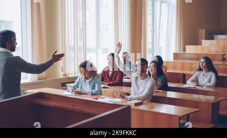 Smart Guy erfolgreicher Student hebt die Hand und spricht mit Professor, während Kommilitonen ihnen zuhören und lächeln. Schüler und Lehrer Beziehungen Konzept. Stockfoto