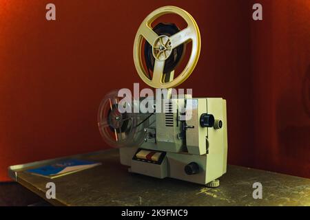 Die Anfänge der Kinematographie. Erstaunliche Entwicklung in der Filmindustrie. Echter antiker weißer Filmprojektor, der auf einem Tisch steht. Roter Hintergrund. Hochwertige Fotos Stockfoto