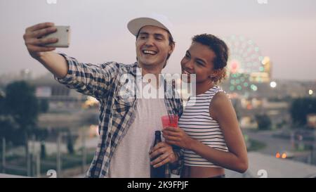 Fröhliches multiethnisches Paar kaukasischer junger Mann und afroamerikanisches Mädchen machen Selfie auf dem Dach mit dem Smartphone, lachen und halten Getränke mit Stadtlichtern im Hintergrund. Stockfoto