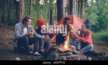 Multirassische Gruppe von Freunden kaukasischen und afroamerikanischen sitzen in Holz um Feuer mit Brille, reden und lächeln, helle Flamme bewegt sich. Stockfoto