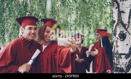 Glückliche junge Menschen Mädchen und Kerl nehmen Selfie nach der Abschlussfeier mit Diplomen tragen Kleider und Mortarboards. Fotografien, Jugend- und Bildungskonzept. Stockfoto