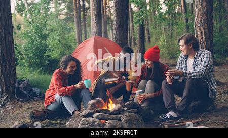 Ein gutaussehender afroamerikanischer Mann gießt heißes Getränk aus der Thermosflasche in Gläser und Tassen, die mit glücklichen, multiethnischen Freunden um das Feuer im Lager sitzen. Stockfoto