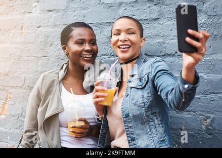 Teilen Sie jeden Moment, während Sie mitgehen. Zwei junge Frauen stehen neben einem Gebäude lächelnd und machen Selfies, während sie ihre kühlen Getränke halten. Stockfoto