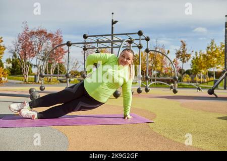 Energisch übergewichtige junge Frau in warmen Sportswear-Training auf Sportplatz im Frühjahr sonnigen Tag. Konzept von Herausforderungen, Träumen, Fitness, gesunder Lebensstil Stockfoto