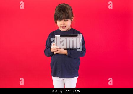 Lächelndes Mädchen mit brünettem Haar, das blaues Sweatshirt und weiße Hosen trägt, das digitale Tablet auf rotem Hintergrund hält und anschaut. Konzept von Call Cente Stockfoto