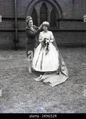 1962, historisch, eine Dame, die die Krone auf dem Kopf eines jungen Teenagers anpasst, das in diesem Jahr zur May Queen gekrönt wurde, während sie in ihrem langen Umzugskleid auf dem Gelände einer Kirche für ein Foto steht, Leeds, England, Großbritannien. Das Maifest ist eine alte Tradition, die den Beginn des Sommers feiert. Stockfoto