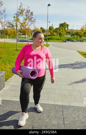 Energisch übergewichtige junge Frau in warmen Sportswear-Training auf Sportplatz im Frühjahr sonnigen Tag. Konzept von Herausforderungen, Träumen, Fitness, gesunder Lebensstil Stockfoto