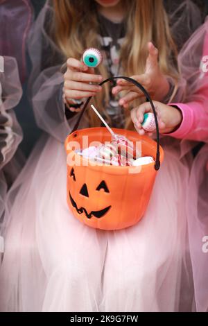 Zugeschnittenes Foto von Mädchen in einer Hexe fancy Kleid hält einen Kürbis-förmigen Süßigkeitenkorb mit gruseligen Gesicht in den Händen. Trick or treat. Verschiedene Süßigkeiten. Konzept von Halloween Familienurlaub, Festival. Stockfoto