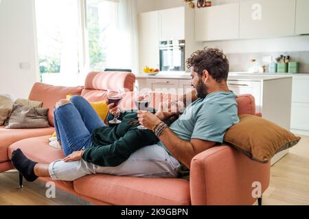 Heterosexuell Romantisches Paar junger Menschen, die sich sorglos auf der Couch entspannen und Wein trinken - die junge Familie hat Spaß beim Kleben auf dem Sofa und genießt es Stockfoto