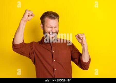 Porträt von sorglosen zufriedenen Mann mit männlichen Bart tragen Burgunder Hemd Schrei ja Klenchfäuste isoliert auf gelbem Hintergrund Stockfoto