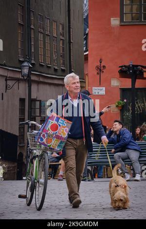 Stockholm, Schweden - 2022. September: Ein alter Mann, der mit Fahrrad und Hund vor den roten und gelben ikonischen Gebäuden am Stortorget in Gamla Stan läuft Stockfoto