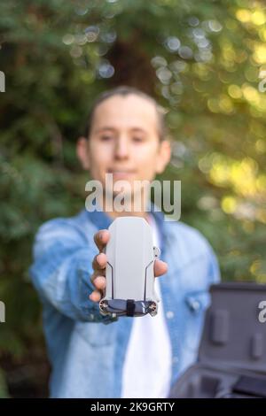 Selektiver Fokus einer kleinen gefalteten Drohne, die von den Händen eines jungen Mannes in Denim-Kleidung im Wald gehalten wird Stockfoto