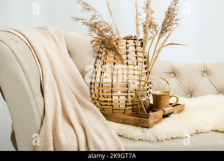 Cozycore oder cottagecore Konzept, warm weich braun beige Interior Design-Objekte. Kuscheliges Wollkaro auf dem Sofa, Kerzenbrenner in Holzlaterne, Teetasse. Stockfoto