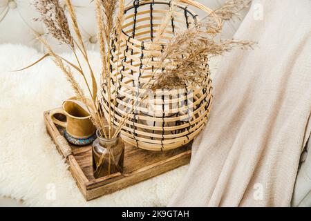 Cozycore oder cottagecore Konzept, warm weich braun beige Interior Design-Objekte. Kuscheliges Wollkaro auf dem Sofa, Kerzenbrenner in Holzlaterne, Teetasse. Stockfoto