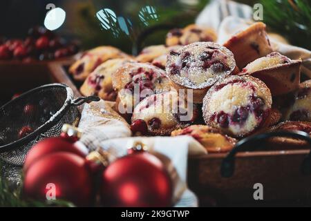 Cranberry-Muffins, die mit Puderzucker bestäubt sind, in einem Tablett mit Weihnachtsbaum und frischen Beeren im Hintergrund. Ornamente und Vintage-Staubwedel in der Nähe. Stockfoto