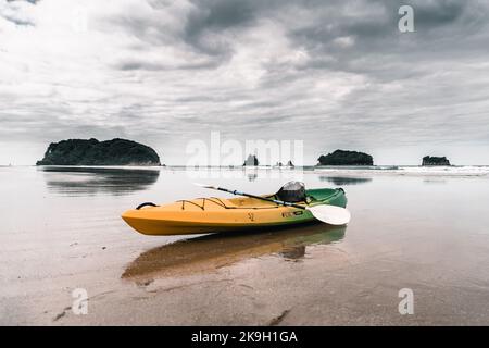 Einsames Kajak mit Paddeln auf der Oberseite am Ufer eines Strandes mit ruhigem Meer und Blick auf Inseln und Wald unter einem trüben grauen Himmel auf Donut Island, neu Stockfoto