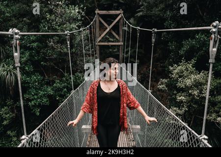 Zerzauste junge kaukasische Frau mit blauen Augen und runden Ohrringen, die ein schwarzes Kleid und eine rote, durchsichtige Jacke auf der Brücke tragen und lächelnd in Richtung Fluss blicken Stockfoto