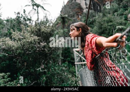 kaukasisches Mädchen mit blauen Augen chaotisch braune Haare mit ihren Händen, die auf dem Metallseil der Waldbrücke in Richtung des Flusses auf der Donut-Insel ruhen Stockfoto