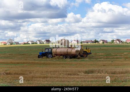 Ein Traktor lädt runde Heuballen auf einen Anhänger in der Mitte eines Feldes, während andere Ballen vor dem Hintergrund von Dorfhäusern liegen. Stockfoto