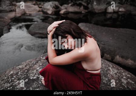 Junge Frau, die auf dem Felsen sitzt, mit den Händen auf dem Kopf Stockfoto