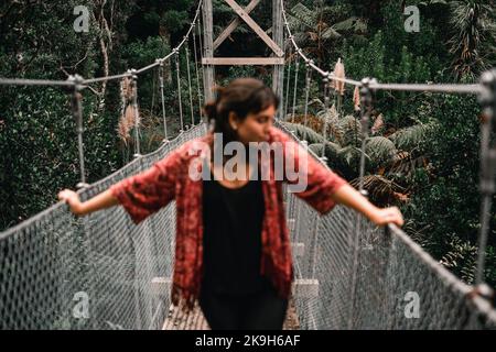 Defokussierte junge kaukasische Frau in roter Jacke und hellen Ohrringen, die sich auf das Geländer einer langen schmalen Holz- und Metallbrücke im Wald lehnen Stockfoto