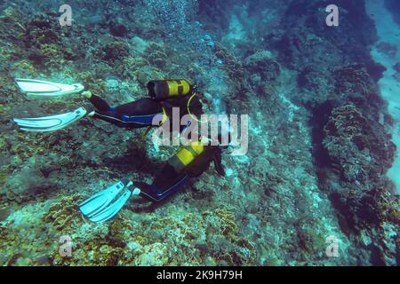 Unterwasserfoto - unbekannter Anfänger Taucher, der mit Korallen über dem Meeresboden schwimmend ist, Tauchlehrer in der Nähe, der seinen Sauerstofftank hält, Blick von oben Stockfoto