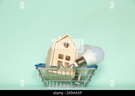 Zwei LED-Glühbirnen und ein kleines Holzhaus liegen in einem Korb aus einem Supermarkt auf einem isolierten Hintergrund, der Preis für Strom, kaufen Licht Stockfoto