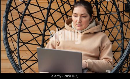 Frau arbeitet auf einem Laptop, der zu Hause im Hängesessel sitzt Stockfoto